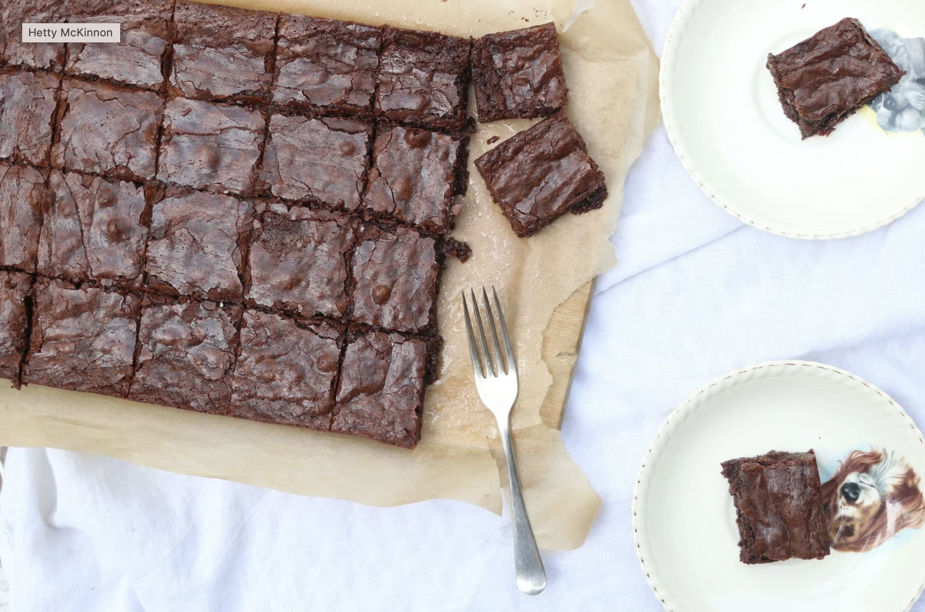 A tray of sliced chocolate brownies on parchment paper with two plates, each holding a brownie, and a fork on the side.