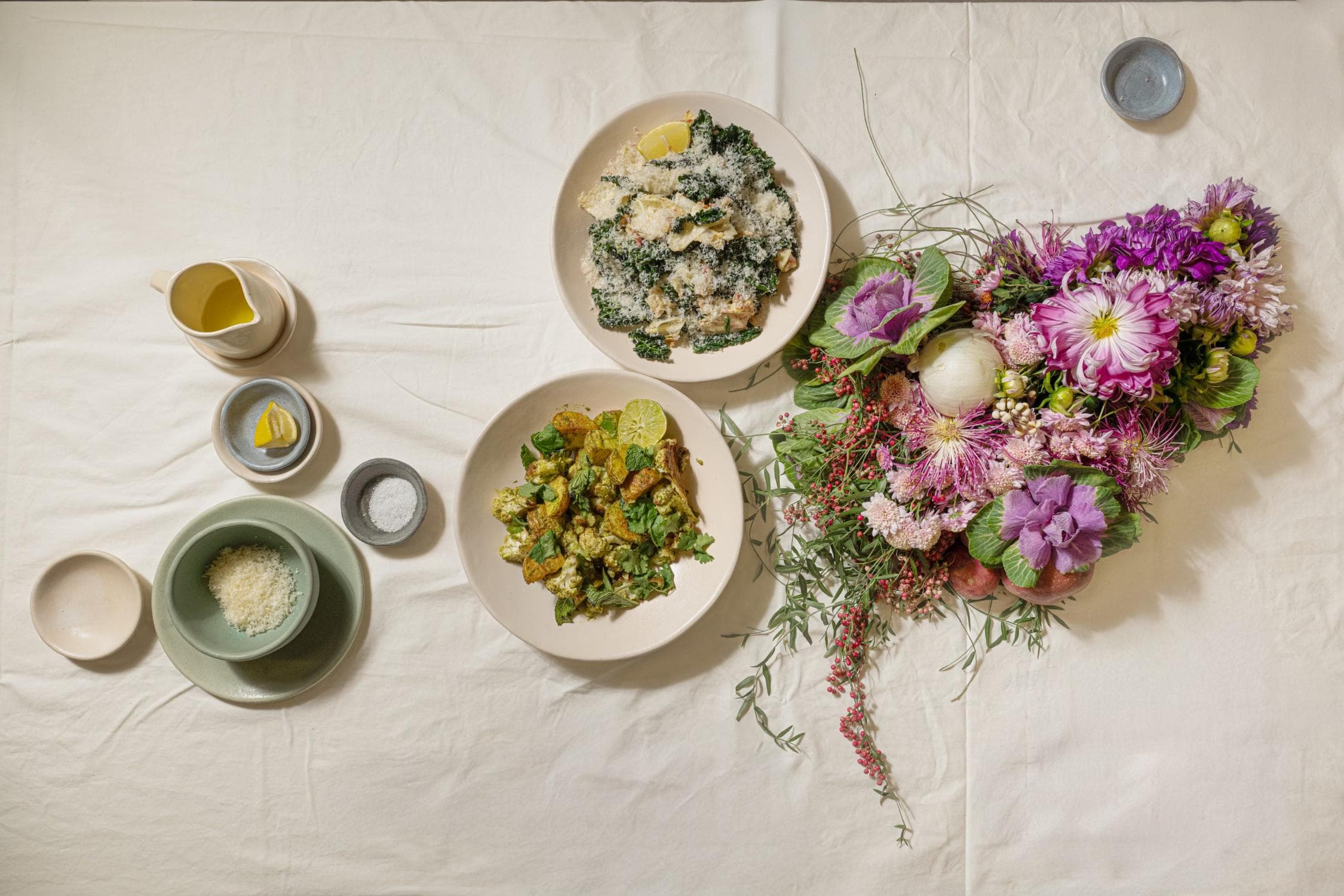 A table set with two plates of food, a pitcher with liquid, a bowl of grated cheese, small bowls of salt and lemon, and a colorful bouquet of assorted flowers.