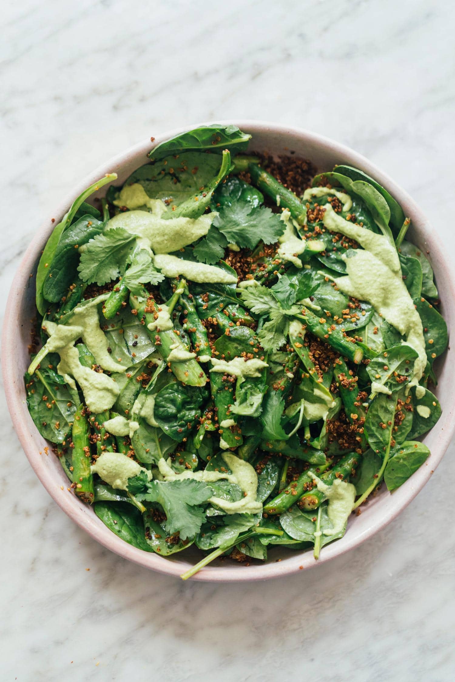 A bowl of leafy green salad with quinoa, fresh herbs, and a light green dressing on a marble surface.