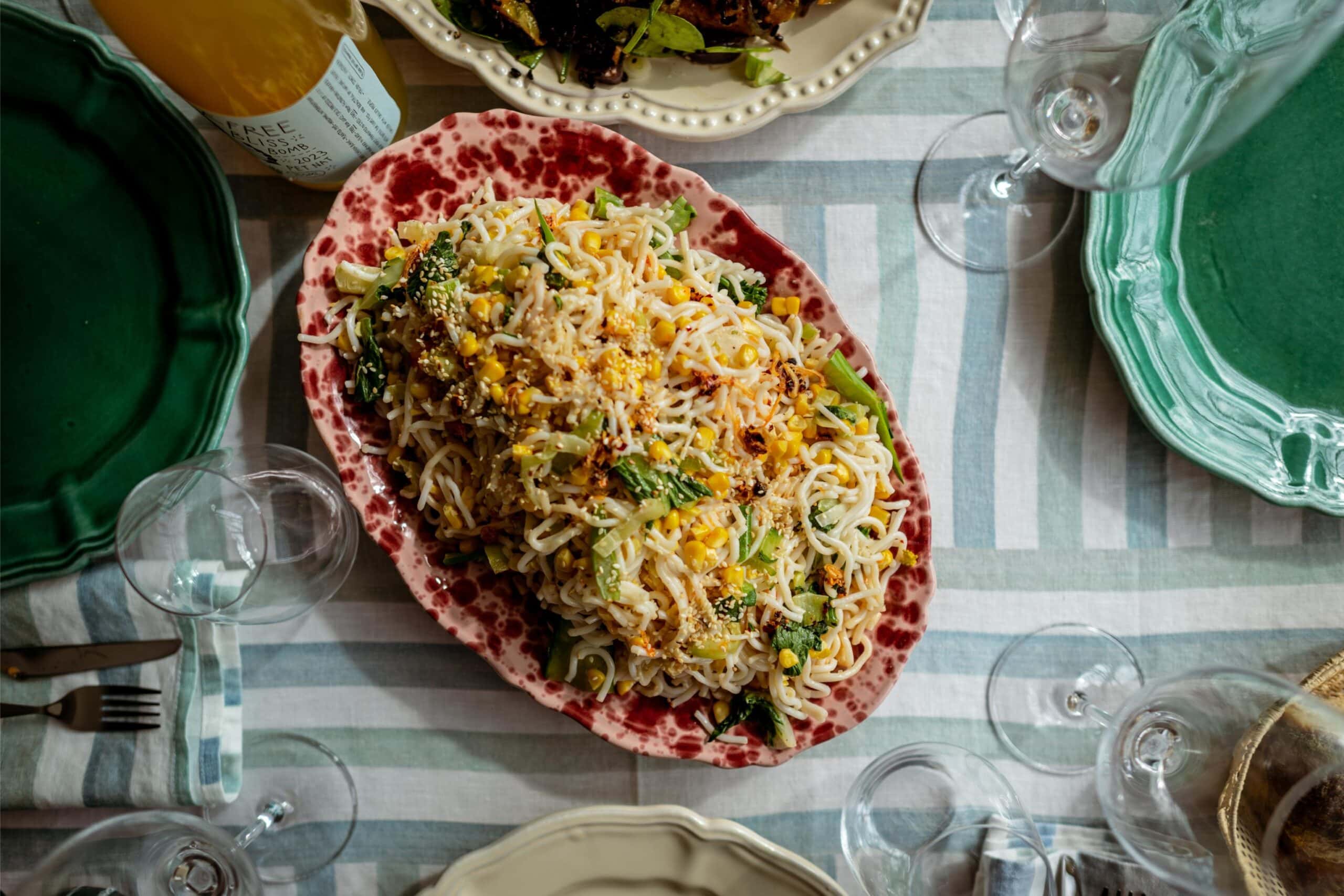 An oval platter of pasta with vegetables and cheese sits on a striped tablecloth, surrounded by empty glasses and green plates.