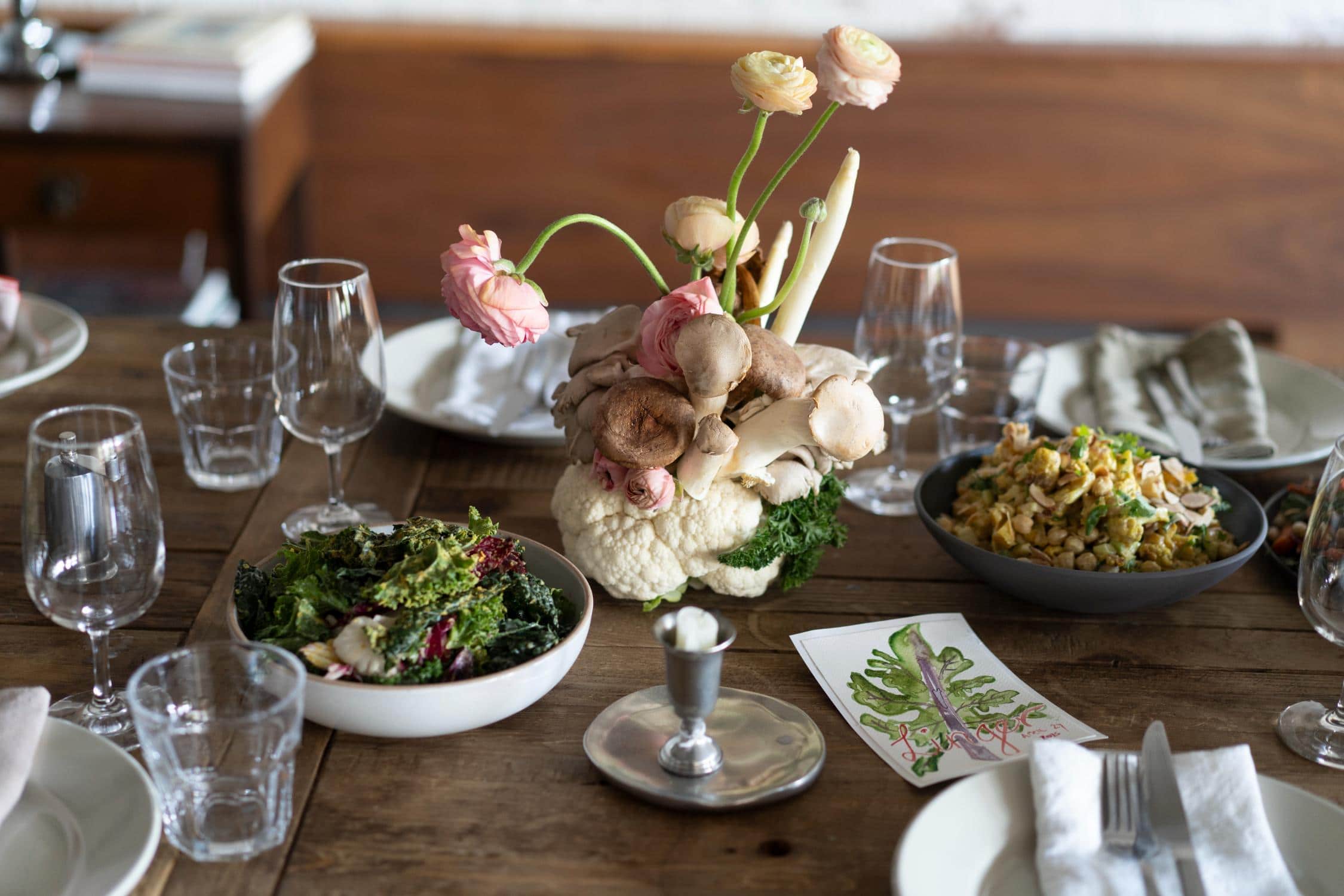 A rustic wooden dining table set with glasses, plates, a salad bowl, a bowl of mixed vegetables, and a floral centerpiece made of cauliflower and mushrooms.