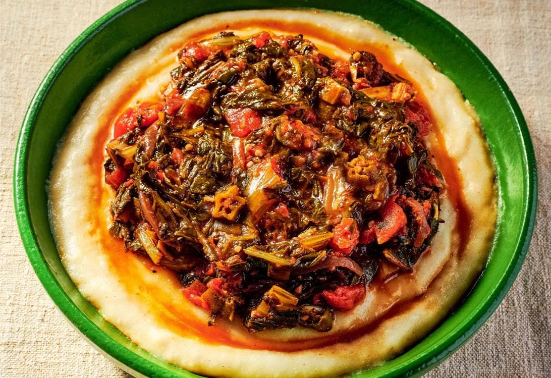 A green bowl filled with a creamy yellow base topped with a tomato and leaf vegetable stew, all set on a beige surface.